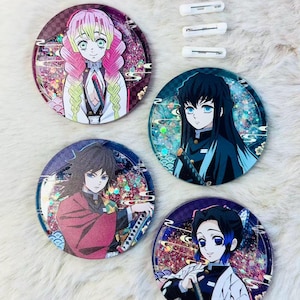 Könnte beinhalten: Vier runde Anime-Charakter-Ansteckbuttons mit Glitzerakzenten, die Charaktere mit rosa, schwarzen und blauen Haaren zeigen. Enthält vier weiße Anstecknadeln. Die Buttons liegen auf einer weißen, flauschigen Oberfläche.