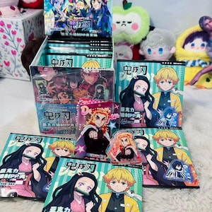 May include: A display of anime-themed trading card packs and acrylic charms. The packaging features colourful illustrations of characters. The cards are in a box, with individual packs and charms scattered around. The text on the packaging is in Japanese.