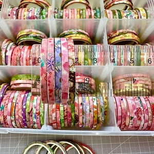 May include: A clear plastic organiser filled with rolls of decorative washi tape. The tape features floral and patterned designs in pink, green, and yellow. The tape rolls are arranged in rows, with a ruler visible for scale. Several loose rolls are at the bottom.