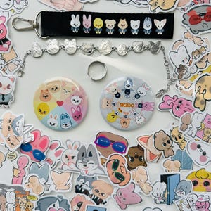 Könnte beinhalten: Eine Sammlung von Aufklebern, ein Armband, ein Ring, ein Schlüsselanhänger und zwei runde Anstecknadeln. Die Aufkleber zeigen verschiedene Cartoon-Tierfiguren in verschiedenen Farben. Das Armband und der Schlüsselanhänger haben kleine tierförmige Anhänger. Die Anstecknadeln haben den Text "SKZOO".