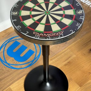 May include: A black and silver dartboard table with a round top featuring a Winmau Diamond Plus dartboard design. The table has a black base and a central support column. The dartboard has red, green, and beige sections.