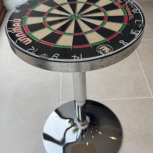 Könnte beinhalten: Runder Barhocker mit Dartscheiben-Oberfläche. Die Dartscheibe hat schwarze, grüne, rote und beige Segmente mit weißen Zahlen. Der Hocker hat ein silbernes Metallgestell und einen kreisförmigen Sockel.