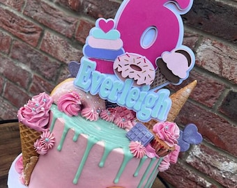 May include: A pink frosted cake decorated with pink frosting swirls, donuts, chocolate bars, and an ice cream cone. The cake is topped with a pink number 6 and the name "Everleigh" in blue.