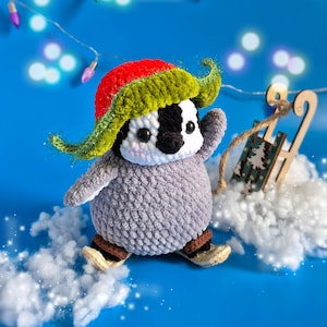 Può includere: Un pinguino all'uncinetto con cappello rosso e verde, viso bianco e nero, corpo grigio e scarpe marroni. Il pinguino è su un letto di soffice bianco con una piccola slitta di legno. Una fila di luci sullo sfondo.