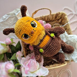 Pu&ograve; includere: Un giocattolo di peluche fatto a mano a forma di ape, con testa gialla, corpo marrone e addome a strisce. L'ape ha grandi occhi espressivi ed &egrave; adornata con piccoli pompon rosa e fiori verdi. Riposa in un cestino di vimini con fiori rosa.