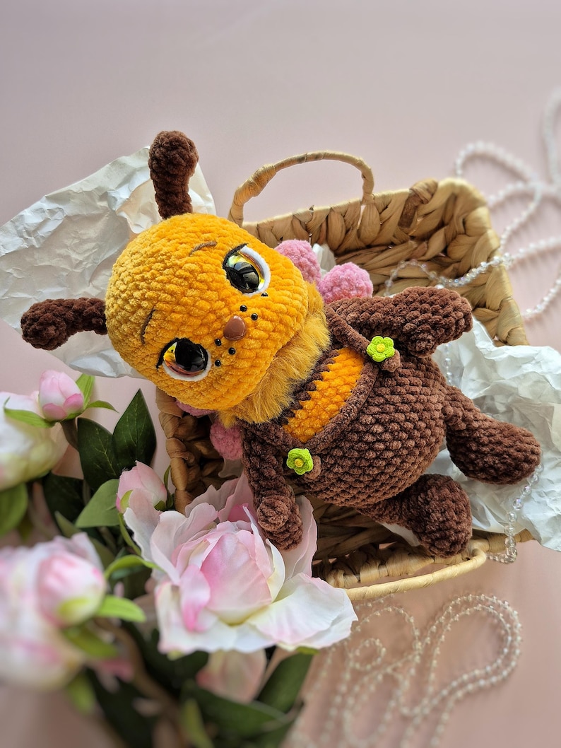 Pu&ograve; includere: Un giocattolo di peluche fatto a mano a forma di ape, con testa gialla, corpo marrone e addome a strisce. L'ape ha grandi occhi espressivi ed &egrave; adornata con piccoli pompon rosa e fiori verdi. Riposa in un cestino di vimini con fiori rosa.
