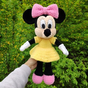 May include: A handmade crochet doll of a cartoon mouse character. The doll has black ears, a pink bow, and a yellow dress. The doll's face is beige with black eyes and a black nose. The doll's feet are pink, and the arms are black with white gloves.