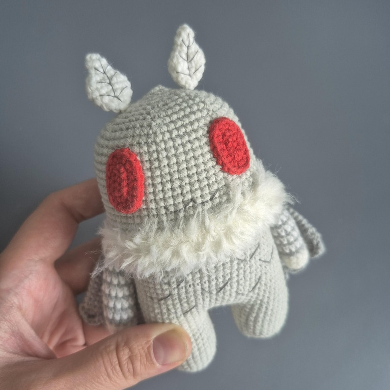 May include: A handmade crochet moth toy in shades of gray, white, and red. The plushie features red eyes, white leaf-shaped antennae, and a fluffy white collar. The toy is held in a hand against a gray background.