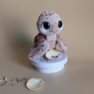 May include: A crocheted turtle toy, predominantly beige and brown, with large, shiny eyes and a gentle expression. It is displayed on a white stand with a seashell and pebbles. The toy has a textured finish, and evokes a seaside theme.