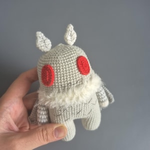 May include: A handmade crochet moth plushie in shades of gray, with red eyes and white accents. The plushie has a fluffy white collar and leaf-shaped details on its head. A unique, whimsical toy.