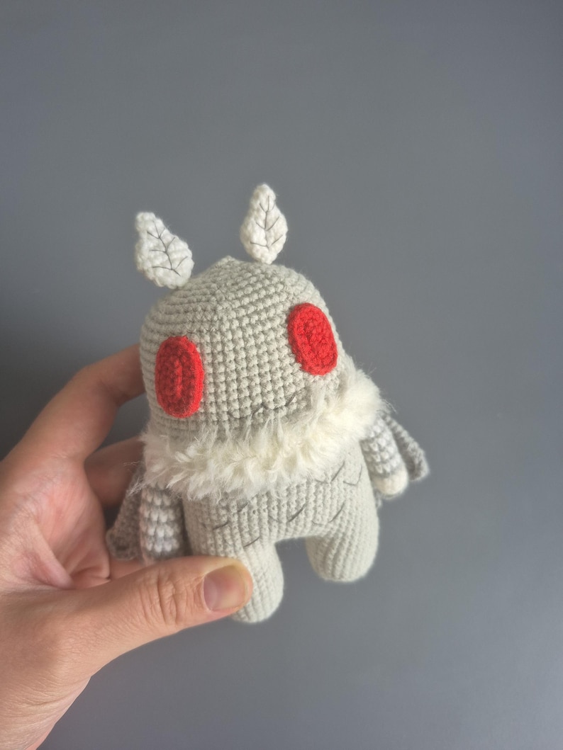 May include: A handmade crochet moth plushie in shades of gray, with red eyes and white accents. The plushie has a fluffy white collar and leaf-shaped details on its head. A unique, whimsical toy.