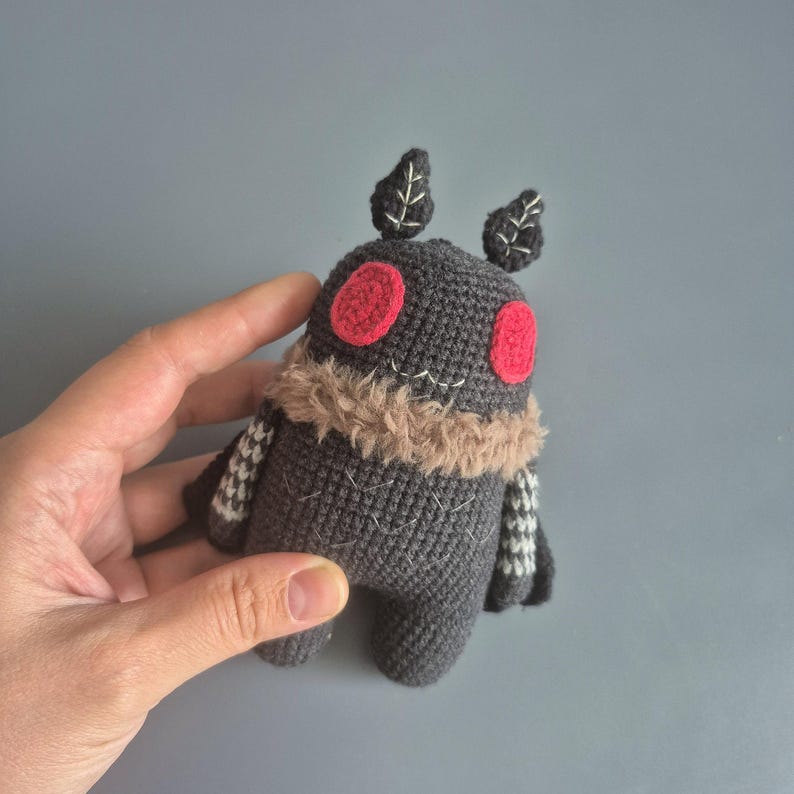 May include: A handmade, dark gray crochet moth plushie with red eyes and a tan fur collar. The moth has small leaf-shaped antennae and patterned wings. The plushie is held in a hand against a gray background.
