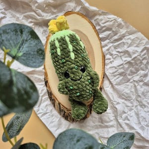 May include: A handmade green crochet cactus plushie with a smiling face, small arms, and beaded accents. The cactus has a yellow flower on top and is displayed on a wooden slice. The background includes a neutral-toned paper and greenery.