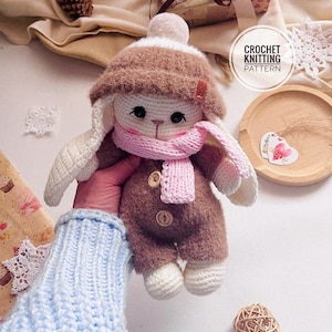 May include: A handmade crochet bunny doll wearing a brown hat, jumpsuit, and pink scarf. The doll has white ears and feet, and is held by a person wearing a blue sweater. The image includes the text "CROCHET KNITTING PATTERN".