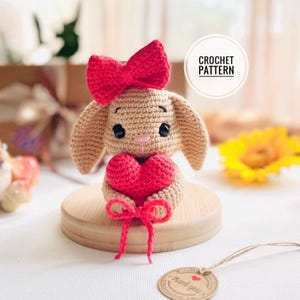 May include: A crocheted bunny figurine with a red bow and a red heart. The bunny is tan with black eyes and a pink nose. The heart is held in its paws and tied with a red bow. The text "CROCHET PATTERN" is in a white circle.