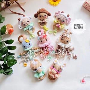 Puede incluir: Colección de juguetes de bebé hechos a mano con ganchillo en varios diseños de animales, incluyendo un cordero, un mono y un caballo. Cada juguete tiene un anillo de madera y está adornado con hilo colorido y detalles florales. Se muestran sobre una superficie blanca.