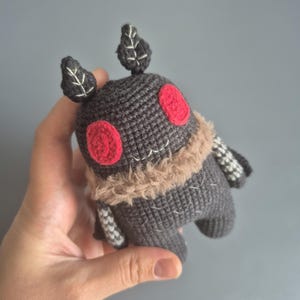May include: A handmade gray crocheted creature with red eyes, a tan fur collar, and leaf-shaped antennae. The plush toy has white stitched details and striped arms. The toy is held in a hand against a gray background.