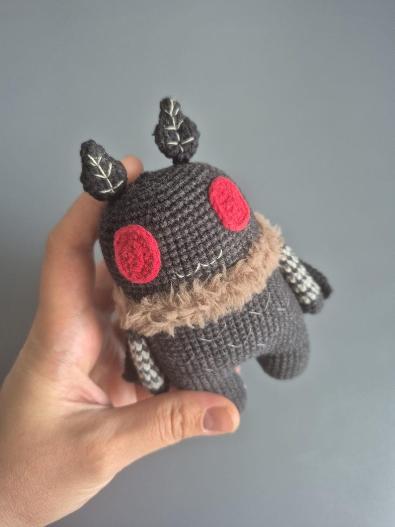 May include: A handmade gray crocheted creature with red eyes, a tan fur collar, and leaf-shaped antennae. The plush toy has white stitched details and striped arms. The toy is held in a hand against a gray background.