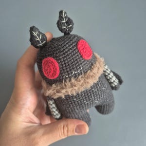 May include: A handmade crochet doll in a whimsical design. The doll features a dark gray body, red eyes, and a tan fur collar. The doll has small leaf-shaped details on its head and patterned arms. A unique and charming handmade toy.