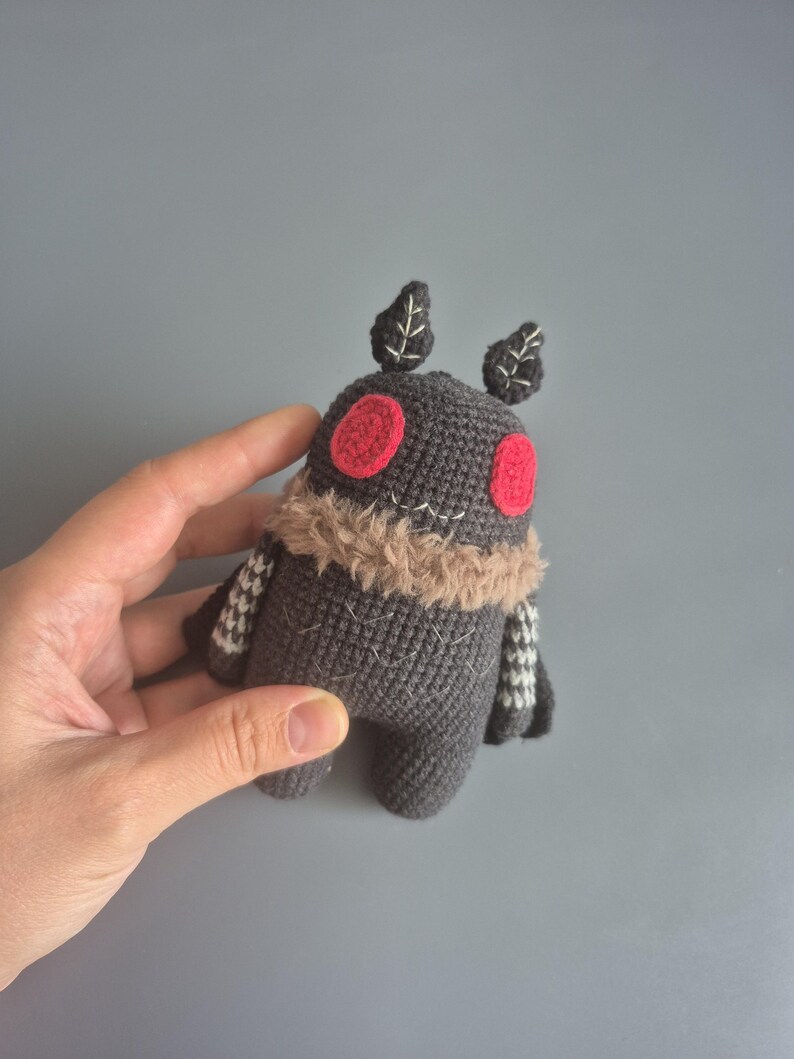 May include: A handmade gray crochet moth toy with red eyes, a tan fur collar, and small leaf-shaped antennae. The moth has small, patterned wings and a stitched design. The toy is held in a hand against a gray background.