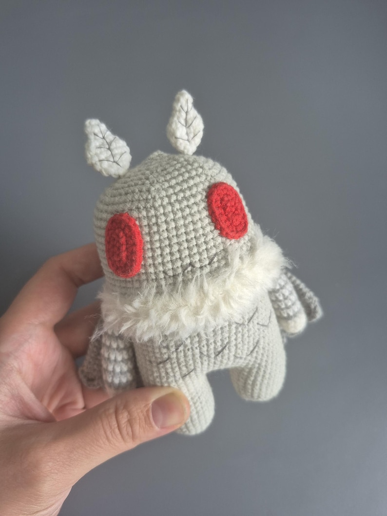May include: A handmade crochet plush toy in the shape of a creature. The toy is primarily light gray with red eyes and white accents. It has leaf-shaped details on its head and a fluffy white collar.