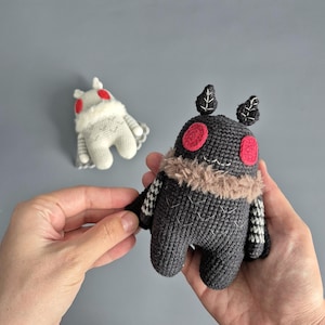 May include: Handmade crochet moth plush toys. One is dark grey with red eyes, a tan collar, and leaf-like antennae. The other is white with red eyes and a white fur collar. Both have small wings and are held in hands.