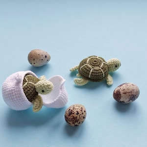 May include: Crocheted turtle emerging from a white eggshell, with another turtle and speckled eggs on a light blue background. The turtles have green and brown shells and light green heads and limbs. The eggs are brown with dark brown spots.