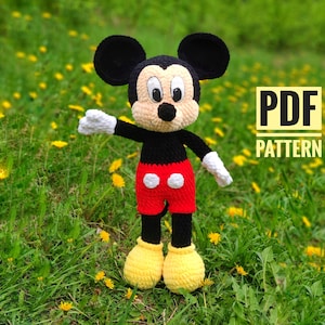 May include: A handmade crochet Mickey Mouse doll, featuring a black head with large ears, a tan face, and a black nose. The doll wears a black body, red shorts with white buttons, white gloves, and yellow shoes. The text "PDF PATTERN" is in a yellow box.