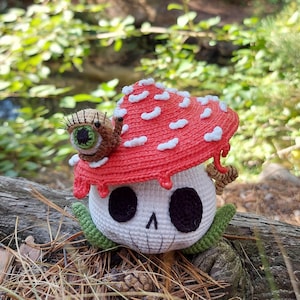 May include: A crocheted mushroom-shaped figure with a skull face, black eye sockets, and a stitched mouth. The red cap has white spots, and a snail with an eye sits on top. Green leaves and a log are in the background.