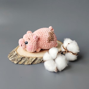 Könnte beinhalten: Ein handgefertigtes rosa gehäkeltes Schwein mit braunen Flecken, das auf einer Holzscheibe liegt. Das Schwein hat kleine Ohren und schwarze Augen. Flauschige weiße Baumwollkugeln sind rechts vom Schwein vor einem grauen Hintergrund angeordnet.
