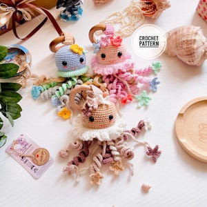 May include: Crocheted jellyfish toys in blue, pink, and tan with wooden rings. Each jellyfish features curly tentacles and decorative flowers or stars. The text "CROCHET PATTERN" is visible.
