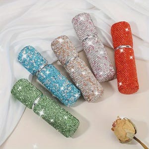 Op de afbeelding: Een assortiment met strass versierde parfumverstuivers in verschillende kleuren, waaronder groen, blauw, brons, roze en rood. Elke verstuiver is bedekt met glinsterende strass steentjes en heeft een zilverkleurige metalen band. De verstuivers zijn gerangschikt op een lichtgekleurd oppervlak.