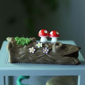 May include: A decorative log with two red and white mushroom figurines, green moss, and small flowers. The log is brown with carved details and sits on a light blue surface. The mushrooms have white stems and red caps with white spots.