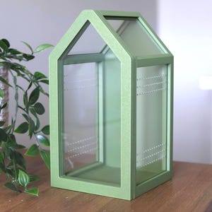 May include: A decorative, house-shaped terrarium with a light green frame and clear glass panels. The terrarium has a pitched roof and is designed to hold small plants or decorative items. The interior features dotted lines for visual interest.