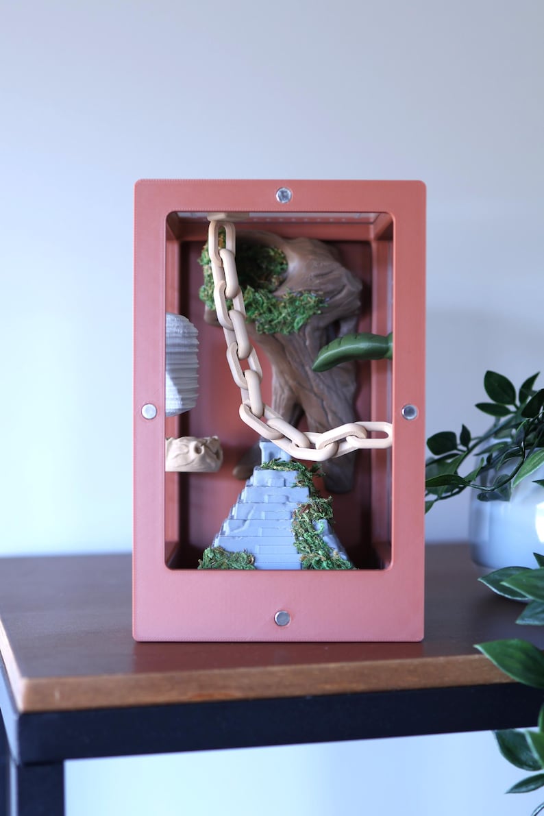 May include: A terrarium with a pink frame, showcasing a miniature landscape. Inside, a light-colored chain hangs over a blue-gray stepped structure covered in green moss. A wooden-like structure and other decorative elements complete the scene.