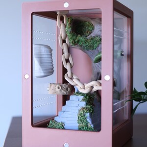 May include: A pink terrarium with a clear front panel, showcasing a miniature landscape. Inside, a light-colored chain hangs, leading to a blue staircase with green moss. A decorative piece and a stack of white objects are also visible.