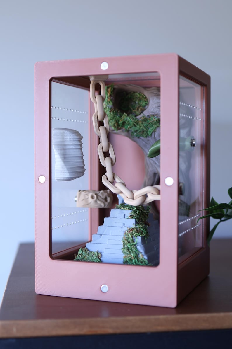 May include: A pink terrarium with a clear front panel, showcasing a miniature landscape. Inside, a light-colored chain hangs, leading to a blue staircase with green moss. A decorative piece and a stack of white objects are also visible.