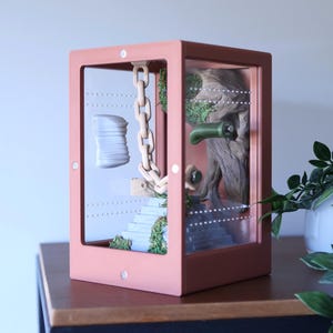 May include: A terrarium with a pink frame, showcasing a miniature scene. Inside, a wooden chain hangs near a stack of white objects and a staircase with greenery. A tree trunk and green accents complete the display.
