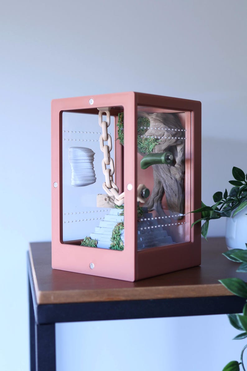 May include: A terrarium with a pink frame, showcasing a miniature scene. Inside, a wooden chain hangs near a stack of white objects and a staircase with greenery. A tree trunk and green accents complete the display.