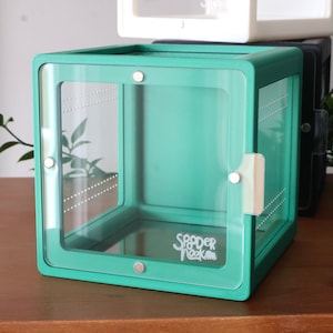 May include: A teal-colored terrarium with clear acrylic panels and a hinged door. The terrarium has a square shape and a white latch. The text "Spider Nook" is visible on the front panel. Other terrariums are stacked behind it.