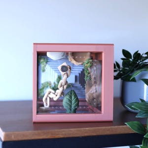 May include: A pink framed shadow box with a miniature scene inside. The scene includes a wooden figure, a green leaf, and a stone structure. The background features a staircase and greenery. The box is displayed on a dark wooden surface.