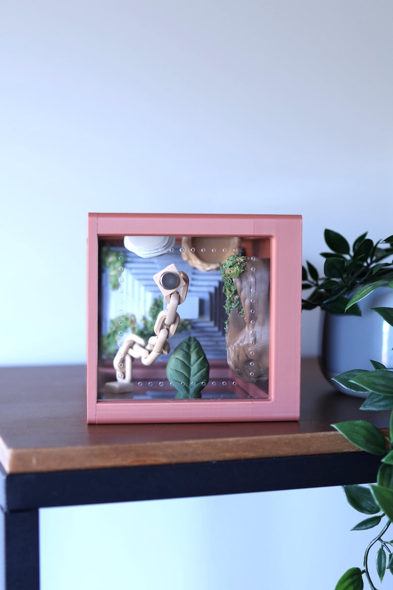 May include: A pink framed shadow box with a miniature scene inside. The scene includes a wooden figure, a green leaf, and a stone structure. The background features a staircase and greenery. The box is displayed on a dark wooden surface.