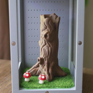 May include: A brown tree stump-shaped planter with detailed bark texture, displayed inside a gray display case. Two miniature red and white mushroom figurines sit on the green artificial grass base, creating a whimsical forest scene.