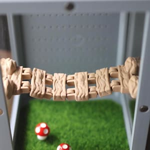May include: A miniature terrarium scene featuring a beige bridge suspended above a bed of green artificial grass. Two red and white mushroom figurines are placed on the grass. The terrarium is enclosed in a gray frame.