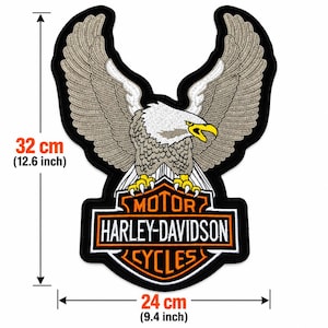 Peut inclure: Un grand écusson brodé représentant un pygargue à tête blanche aux ailes déployées perché sur un logo Harley-Davidson Motor Cycles. L'écusson mesure 32 cm de hauteur et 24 cm de largeur, avec des fils de couleurs noire, blanche, grise et jaune.