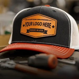 以下が含まれることがあります： 赤いつばの黒と白のトラッカーハット。帽子には「YOUR LOGO HERE」と「MADE IN USA」の文字が入った革製のパッチが付いています。帽子はメッシュの背面と湾曲したつばが付いています。