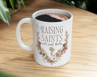 May include: White ceramic coffee mug with the text "RAISING SAINTS not just kids" in a decorative font, surrounded by a floral wreath and a cross. The mug is filled with dark coffee.
