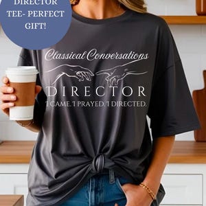 Pode incluir: Uma camiseta cinza com um gráfico branco de duas mãos que se alcançam. O texto "Classical Conversations" está acima das mãos e o texto "DIRECTOR I CAME. I PRAYED. I DIRECTED." está abaixo das mãos.
