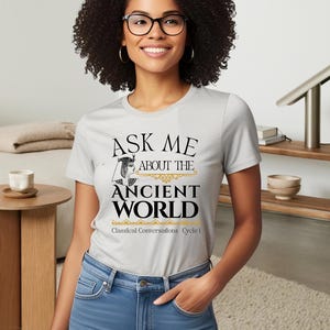May include: A gray t-shirt with the text "Ask me about the Ancient World" in black and gold. The text is surrounded by a decorative border. Below the text is "Classical Conversations Cycle I" in black.