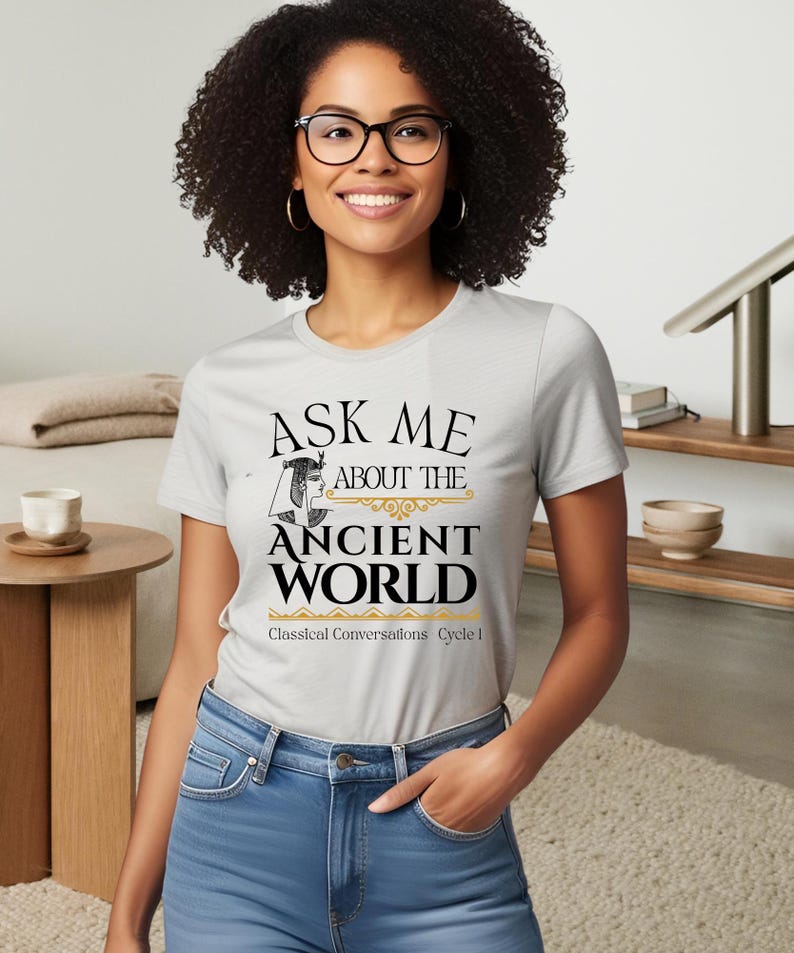 May include: A gray t-shirt with the text "Ask me about the Ancient World" in black and gold. The text is surrounded by a decorative border. Below the text is "Classical Conversations Cycle I" in black.